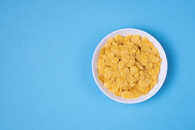 Cornflakes in white bowl on blue background for cereal healthy food. Top view. Copy, empty space for text