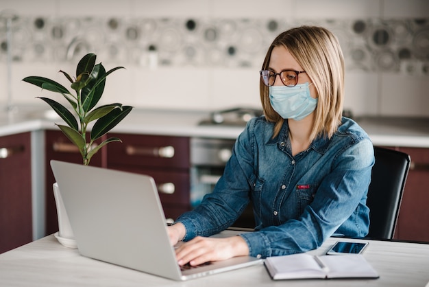 Coronavirus. Business woman working in home, wearing protective mask in quarantine. Stay at home. Girl learns, using laptop computer in home office. Freelance. Communication and technology concept.