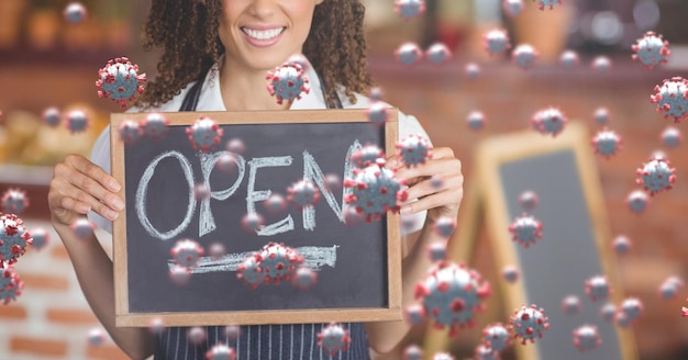 Photo coronavirus cells floating over mixed race woman chef holding an open sign, smiling, restaurant in the background. coronavirus and restaurant business.