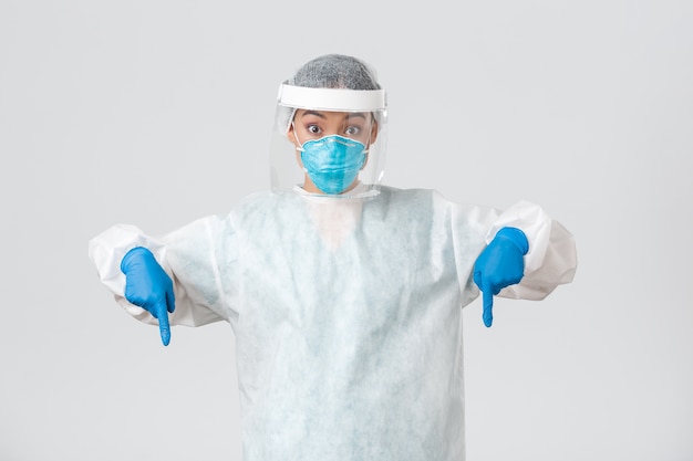 Photo , coronavirus disease, healthcare workers concept. surprised and excited female doctor, physician in personal protective equipment pointing fingers down, standing white wall.