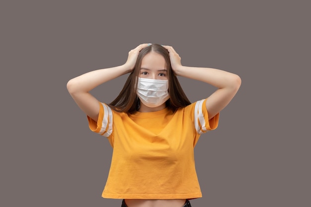 Coronavirus epidemic and panic people's fear of getting sick Young girl in protective medical mask holds her head scared isolated on gray background