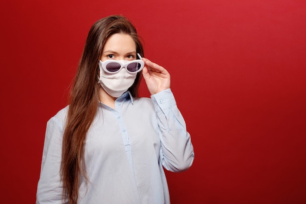 Coronavirus pandemic close-up portrait of young woman on red  in protective medical mask and sunglasses