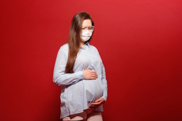 Coronavirus pandemic, young pregnant woman on red background in protective medical mask holds on stomach