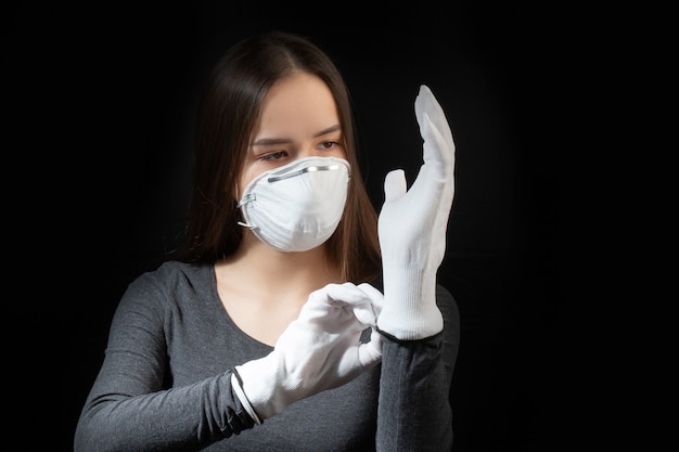 Coronavirus protection. Young woman in a white antibacterial medical mask puts on gloves on a black wall