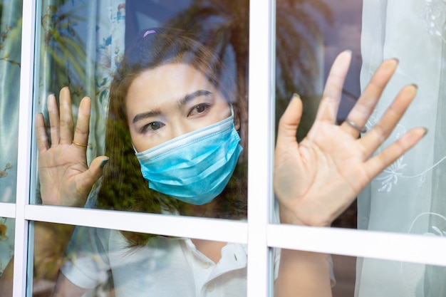 Coronavirus. Sick woman of corona virus looking through the window and wearing mask protection and recovery from the illness in home. Quarantine. Patient isolated to prevent infection new normal.