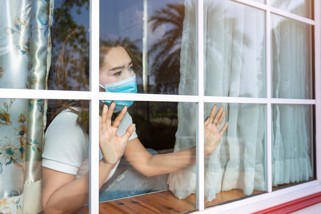 Coronavirus. Sick woman of corona virus looking through the window and wearing mask protection and recovery from the illness in home. Quarantine. Patient isolated to prevent infection new normal.