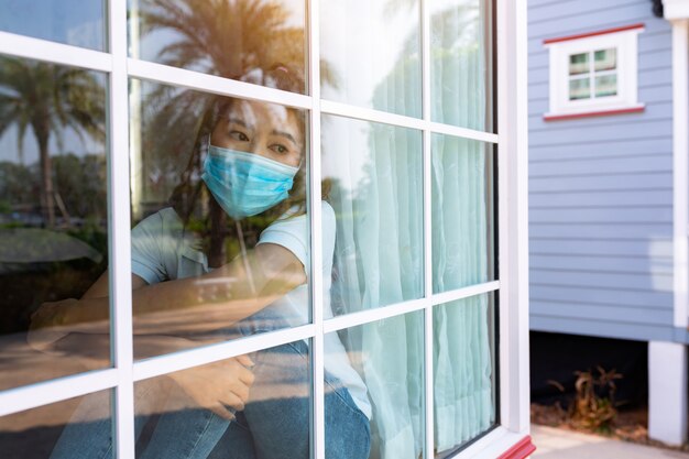 Coronavirus. Sick woman of corona virus looking through the window and wearing mask protection and recovery from the illness in home. Quarantine. Patient isolated to prevent infection new normal.