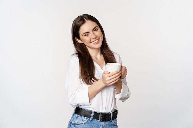 Corporate woman standing with coffee tea mug and smiling drinking from cup standing happy against wh...