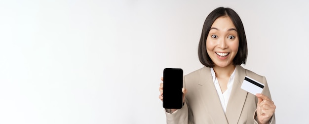 Corporate woman with happy enthusiastic face showing credit card and smartphone app screen standing in suit over white background