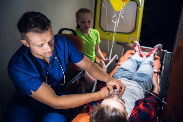 A corpsman in a uniform applying an oxygen mask to an unconscious woman lying on a stretcher in an ambulance car.