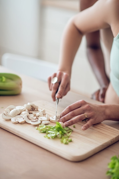 Correct breakfast. Neat slender hands of caucasian woman chopping mushrooms and greens on kitchen surface, faces are not visible