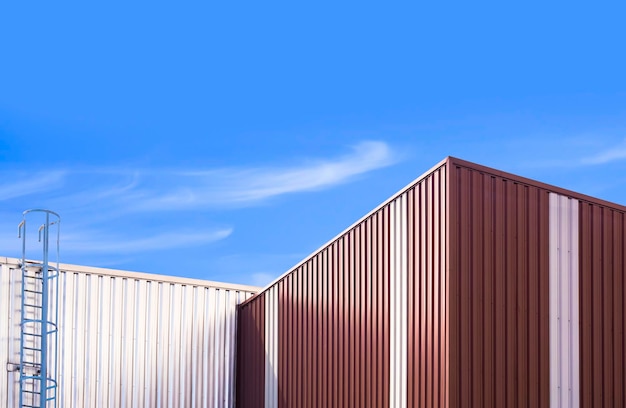 Corrugated steel wall of 2 warehouse buildings with metal stair against blue sky background