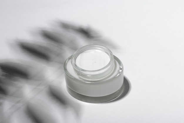 Cosmetic cream or mask in a glass jar with hard shadows from the leaves on a white background. Natural organic care for face and body skin. View from above.