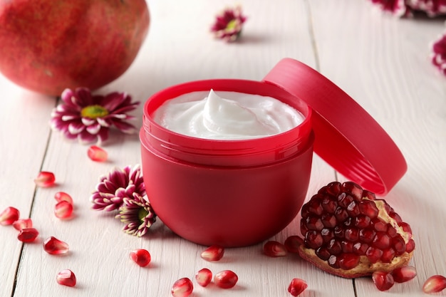 Cosmetic cream in a red jar with flowers and fresh pomegranate on a white wooden table.