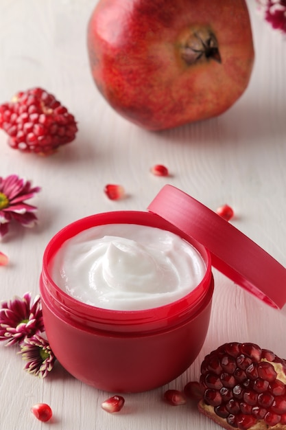 Cosmetic cream in a red jar with flowers and fresh pomegranate on a white wooden table.