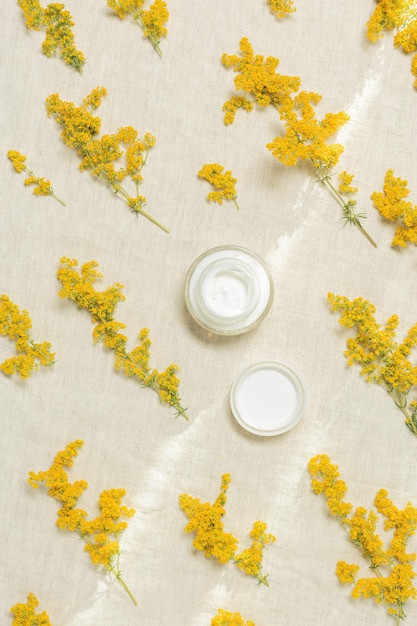 Cosmetic cream in round container and yellow bedstraw herb wildflowers on natural textile