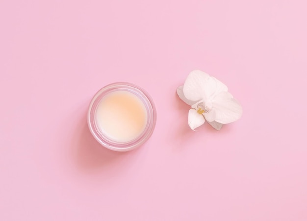 Cosmetic glass jar near white orchid flower on light pink top view