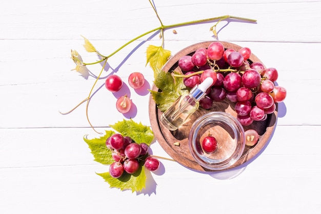 Cosmetic oil whey based on grape seed oil in a glass vial with a pipette on a wooden tray with a bunch of grapes top view white wooden background