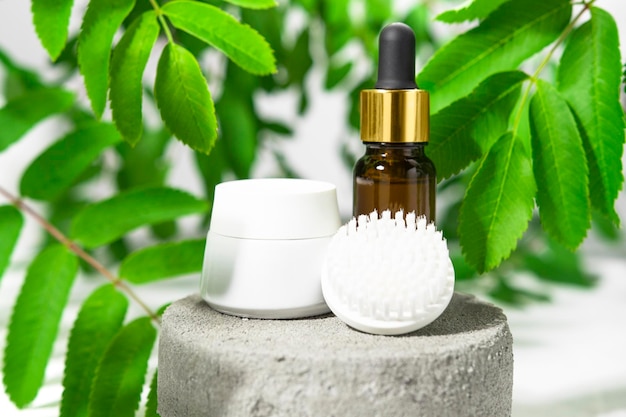 Cosmetic products on a gray stone podium with plants body care cream with brush and serum front view