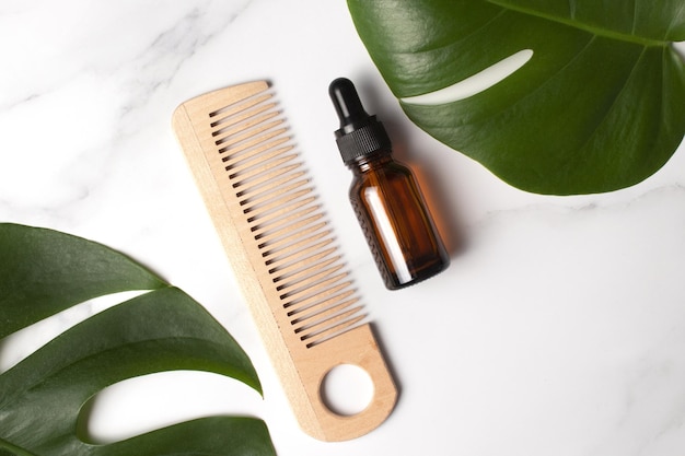 Cosmetic serum in brown glass bottle and wooden hair comb on a marble background with monstera palm leaf Natural cosmetics concept Top view flat lay copy space