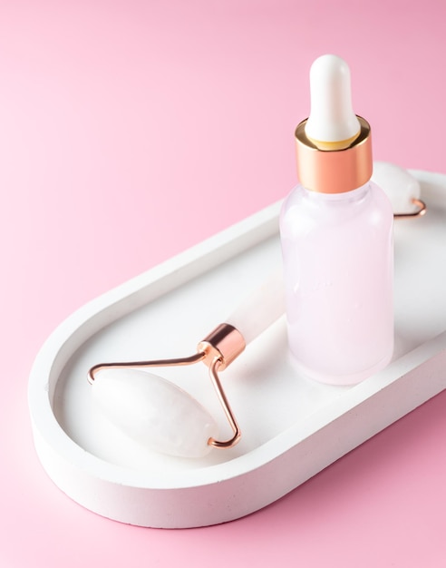 Cosmetic serum in glass bottle and face roller on white concrete tray on pink background. Skincare, natural organic cosmetics concept.