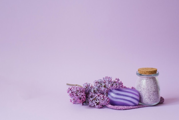 Cosmetic spa set for body care Sea salt in glass jar organic soap washcloth glove and lilac flowers on purple background with copy space