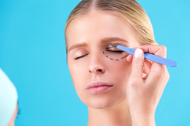 Cosmetic surgeon examining female client in office. Doctor draws lines with a marker, the eyelid before plastic surgery, blepharoplasty. Surgeon or beautician hands touching woman face. Rhinoplasty