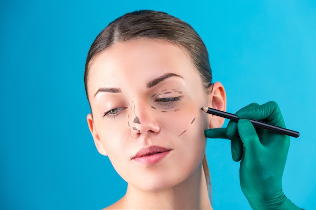 Cosmetic surgeon examining female client in office. Doctor draws lines with a marker, the eyelid before plastic surgery, blepharoplasty. Surgeon or beautician hands touching woman face. Rhinoplasty