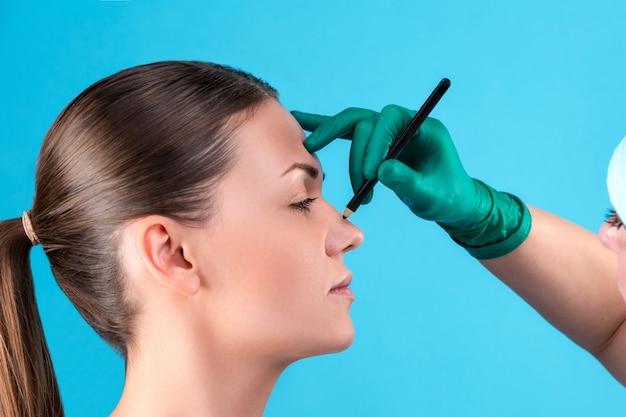 Cosmetic surgeon examining female client in office. Doctor draws lines with a marker, the eyelid before plastic surgery, blepharoplasty. Surgeon or beautician hands touching woman face. Rhinoplasty