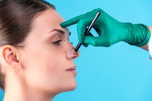 Cosmetic surgeon examining female client in office. Doctor draws lines with a marker, the eyelid before plastic surgery, blepharoplasty. Surgeon or beautician hands touching woman face. Rhinoplasty