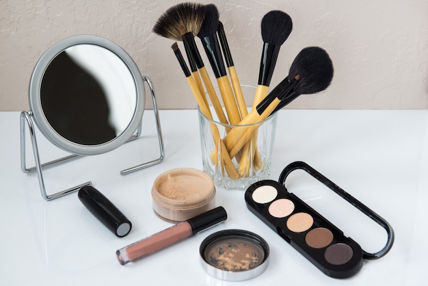 Cosmetics with makeup brushes and mirror on a white table.