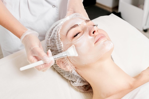 Cosmetologist applies cream mask on woman face for rejuvenation face skin procedure in beauty salon