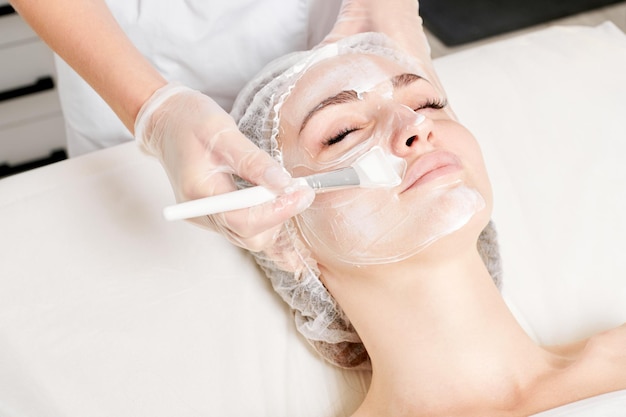 Cosmetologist applies cream mask on woman face for rejuvenation face skin procedure in beauty salon