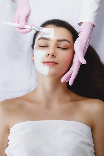 The cosmetologist applies a white clay mask to the face of a beautiful young woman