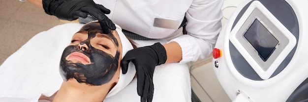 Cosmetologist applying black mask on the face of a beautiful woman for carbon peel