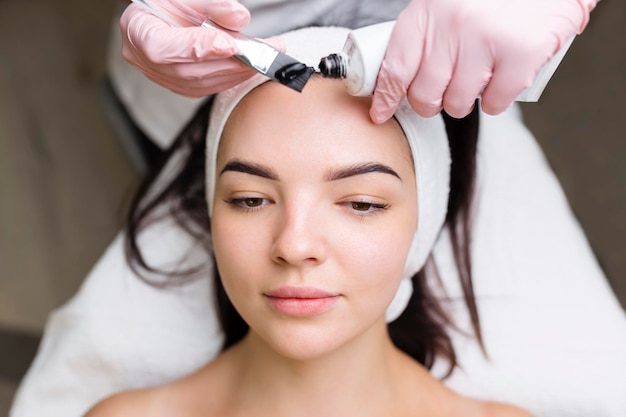 Cosmetologist applying black mask on pretty woman face wearing black gloves gorgeous woman in spa having facial procedures