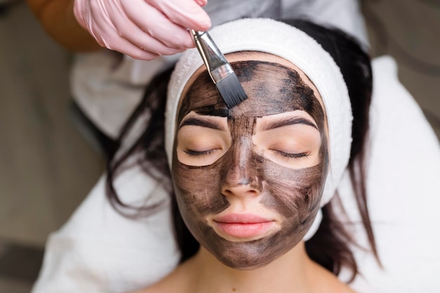 Cosmetologist applying black mask on pretty woman face wearing black gloves gorgeous woman in spa having facial procedures