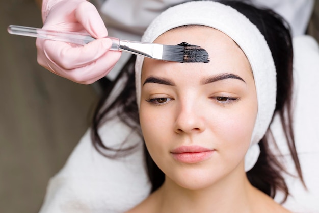 Cosmetologist applying black mask on pretty woman face wearing black gloves gorgeous woman in spa having facial procedures