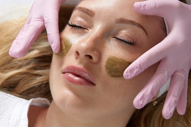 Cosmetologist applying a clay mask to the face of beautiful woman Spa treatment and face care in the beauty salon