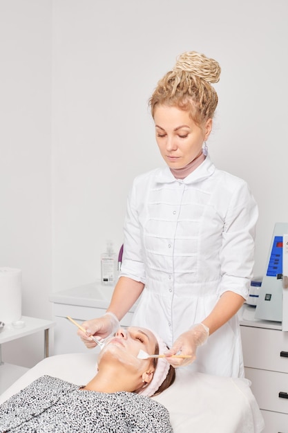 Cosmetologist applying cosmetic cream mask on female face for rejuvenation procedure in beauty salon