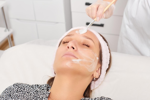 Cosmetologist applying cosmetic cream mask on female face for rejuvenation procedure in beauty salon