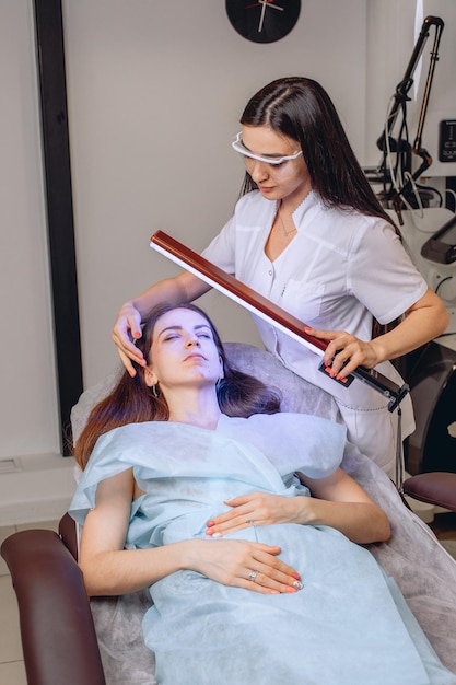 The cosmetologist examines the girl's face on the couch through a purple lamp. Diagnosis facial skin.