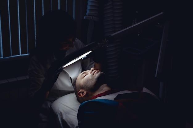 A cosmetologist examines a man's facial skin in the dark under a lamp in preparation for the procedure