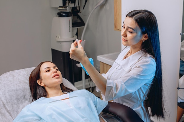 The cosmetologist fills the syringe with a means for carrying out cosmetic services for the girl lying next to her on the couch.