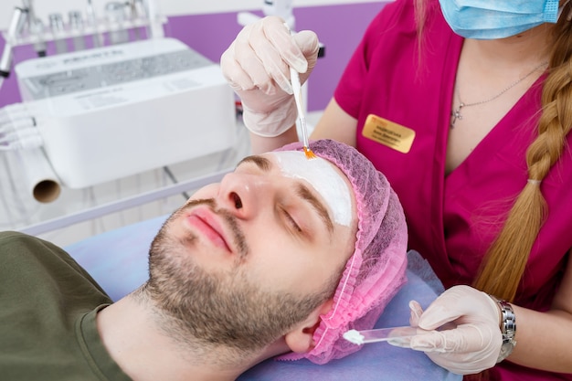 Cosmetologist is applying white moisturizing mask on man's face using brush in beauty clinic, face closeup. Beauty industry concept. Guy is lying on couch. Beautician woman making spa procedure.