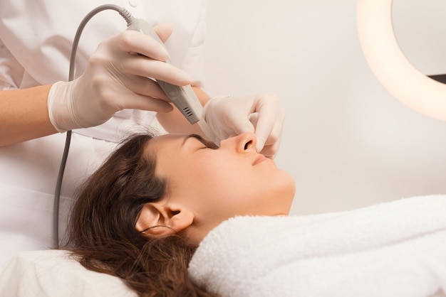 Cosmetologist is cleaning a womans face with the ultrasound spatula.