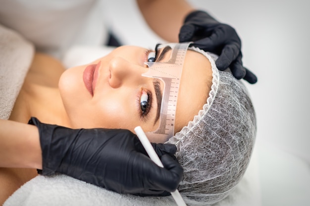 Cosmetologist is measuring with ruler the brows of young woman