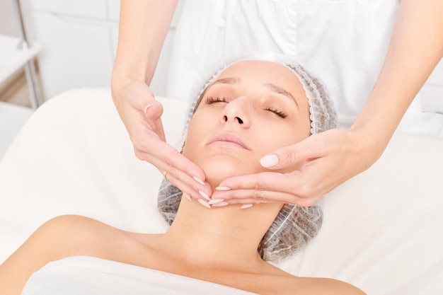 Cosmetologist massages cream mask into woman face skin for rejuvenation procedure in beauty salon