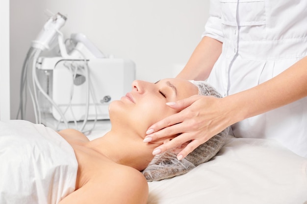 Cosmetologist rubbing moisturizing cream mask into woman face skin for rejuvenation in beauty salon