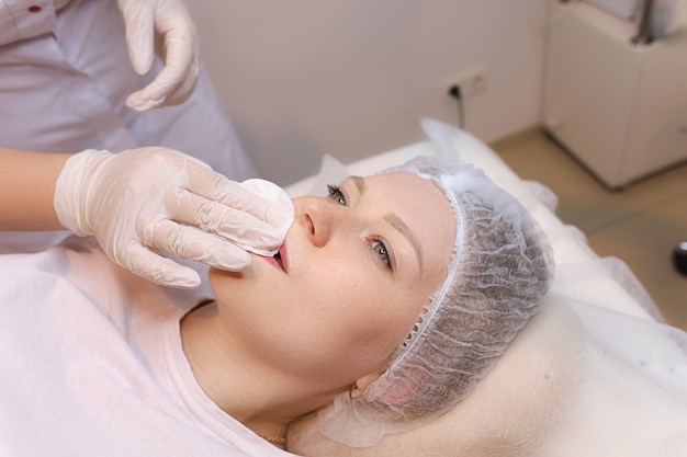 The cosmetologist wipes the remnants of anesthesia from the client's lips with a cotton sponge